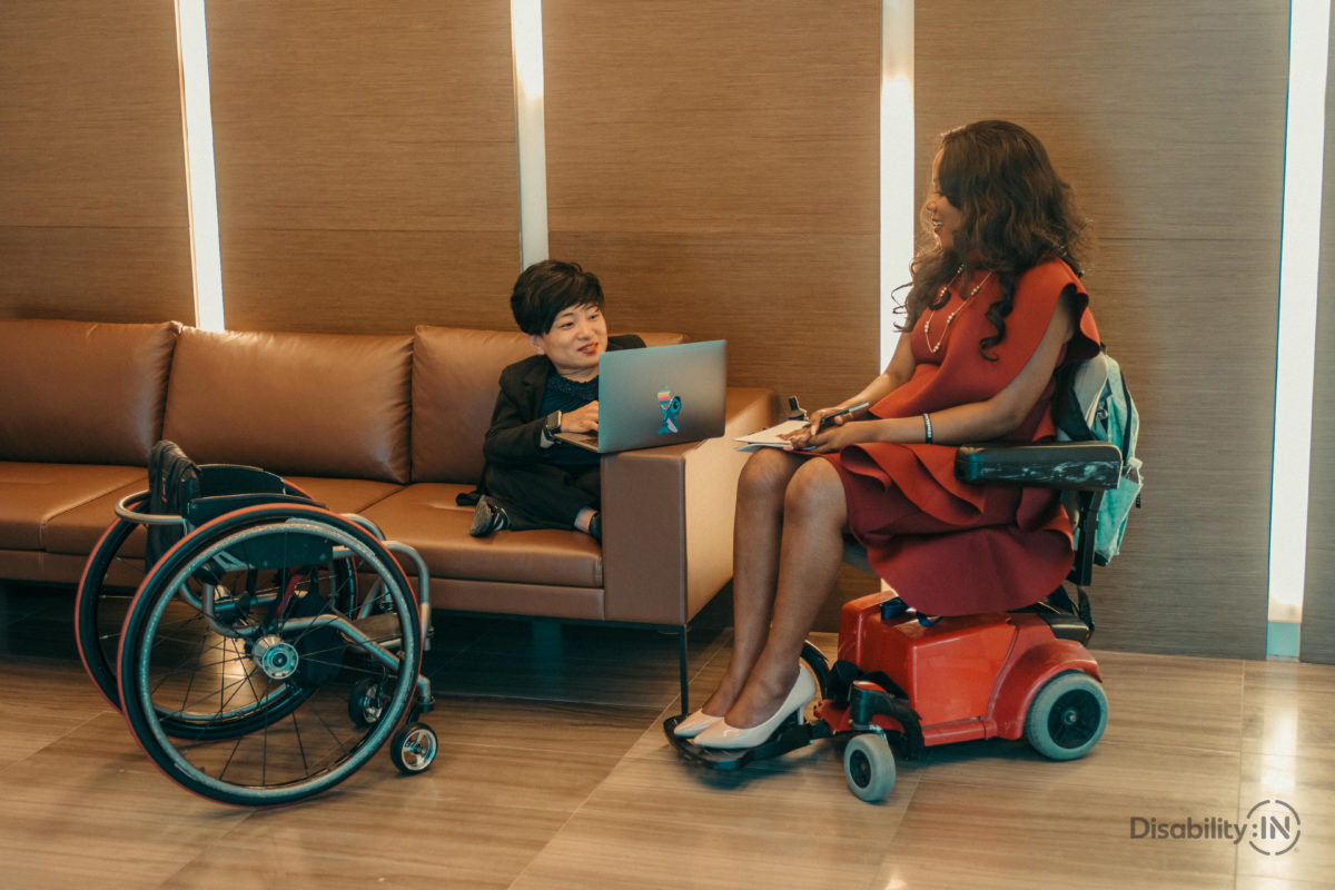 Two women with disabilities, one sitting on a couch with a laptop, enjoying a moment to discuss work related things.